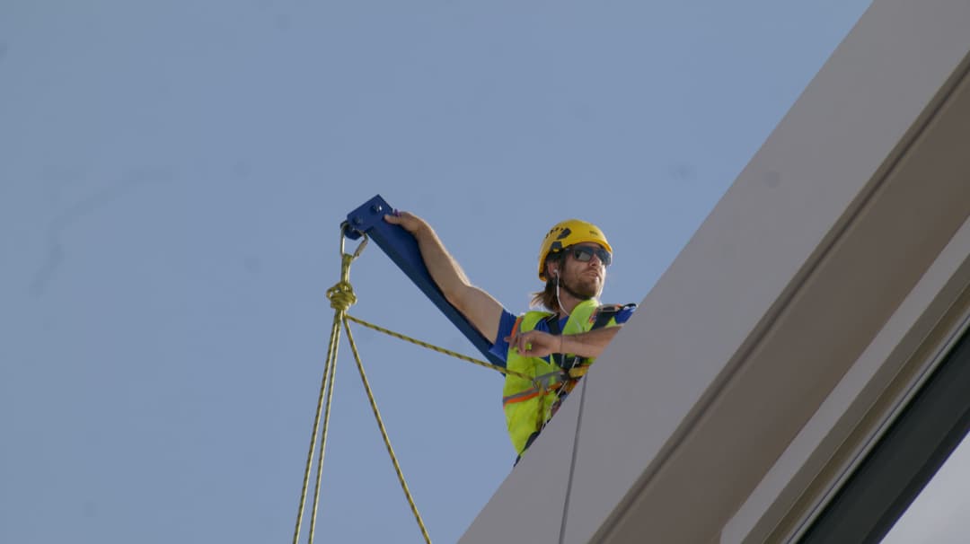 Construction worker in safety gear securing equipment on a building rooftop against a blue sky.