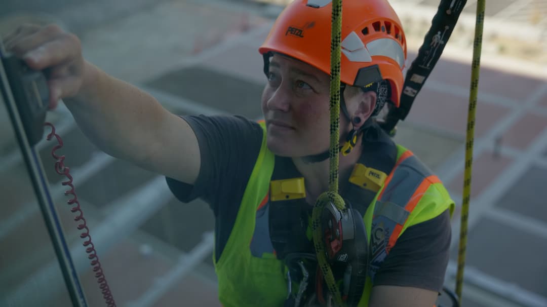 Worker in safety gear cleaning a high-rise window with harness and safety helmet.