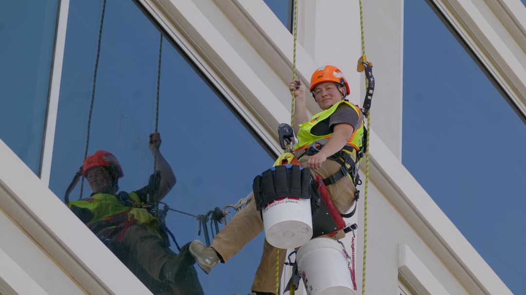 Window cleaner in safety gear working on a high-rise building, reflecting in the glass.