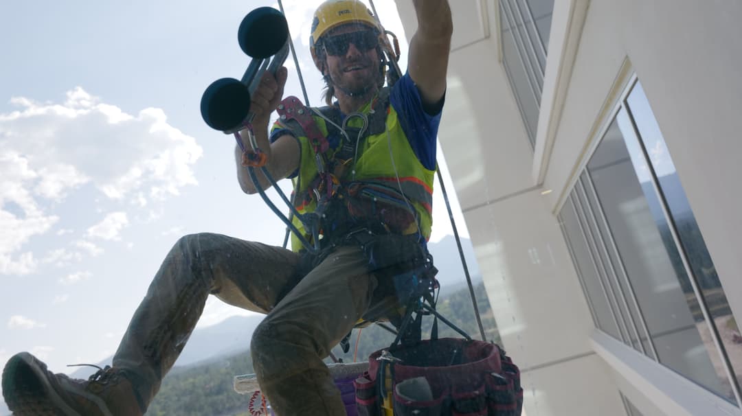 Window cleaner suspended from a building, wearing a helmet and harness, smiling while working.