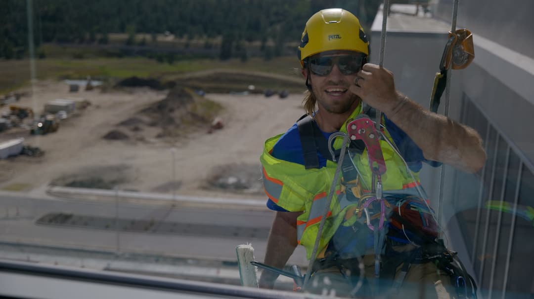 Window cleaner in safety gear on a high-rise, overlooking construction site and landscape.