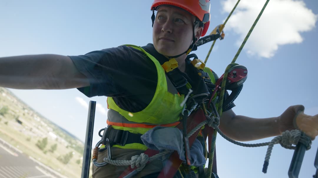 Roofer in safety gear, harnessed and working at height against a clear blue sky.