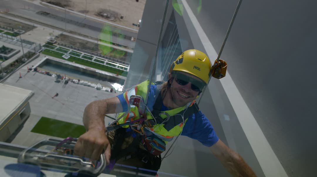 Window cleaner in a hard hat and harness, smiling while suspended from a building.